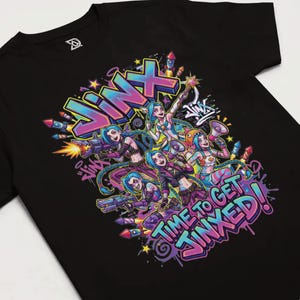 May include: Black t-shirt featuring a colorful graphic of multiple characters with blue hair, holding weapons and a megaphone. The word "Jinx" is prominently displayed, along with the phrase "Time to get Jinxed!" in a graffiti style.