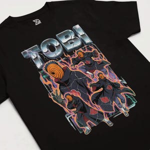 Puede incluir: Camiseta negra con un diseño del personaje Tobi de la serie de anime Naruto. El diseño incluye la palabra "TOBI" en plata con efecto de relámpago e ilustraciones del personaje en acción.