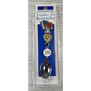 Puede incluir: Una cuchara de recuerdo de plata con un mango de cobre macizo. La cuchara presenta una pequeña bandera estadounidense y un escudo con el texto "Tex Fort Custer Co.". La cuchara está empaquetada en una caja de plástico transparente con el texto "Yours... To Remember" y "FORT Made in USA."