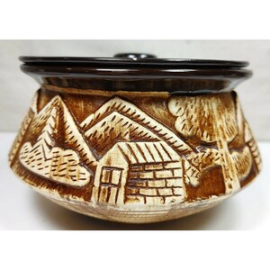 May include: A brown ceramic pot with a lid, featuring a raised design of houses and trees. The pot has a white background with brown details.