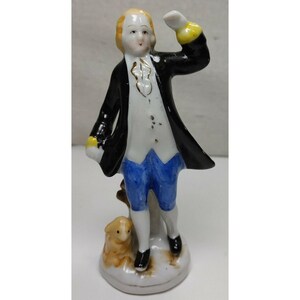 May include: A porcelain figurine of a person wearing a black coat, white vest, and blue breeches, with one arm raised. The figure has blonde hair and stands on a base next to a small, light brown animal, possibly a dog, with painted details.