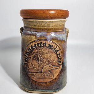 May include: A brown ceramic jar with a wooden lid. The jar has a circular design with the words "Minnesota Wild Rice" and a loon illustration.
