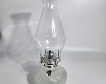 May include: A clear glass oil lamp featuring a bulbous, frosted glass base with a geometric starburst pattern. The lamp includes a silver-toned metal burner assembly and a tall, transparent glass chimney with a crimped top edge.
