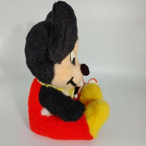 May include: A vintage plush Mickey Mouse toy with black fur, red shorts, and yellow shoes. The toy is sitting with its legs crossed and has a friendly expression.