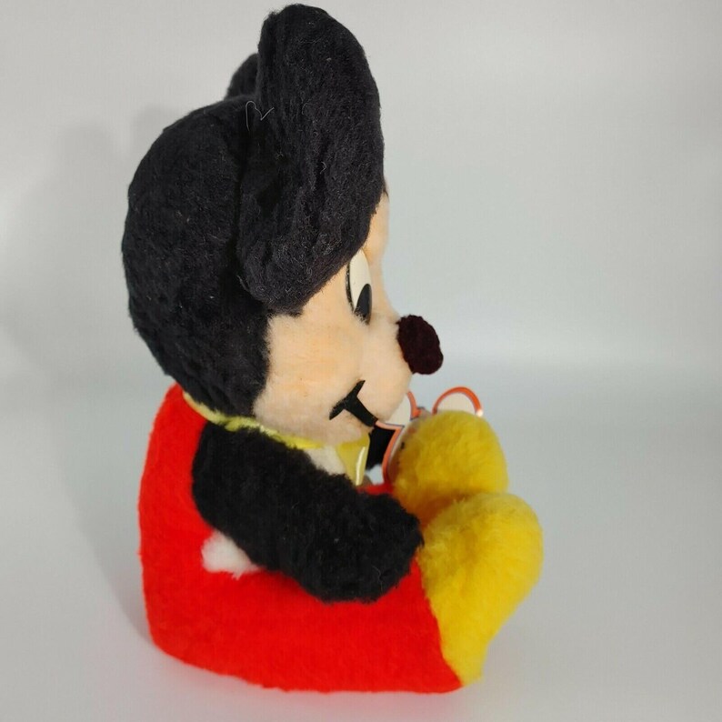May include: A vintage plush Mickey Mouse toy with black fur, red shorts, and yellow shoes. The toy is sitting with its legs crossed and has a friendly expression.