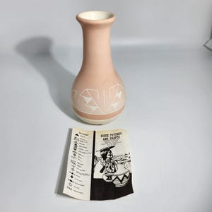 May include: A peach-coloured vase with a white rim and geometric designs. The vase has a slender neck and a rounded body. A brochure with the text "Sioux Pottery and Crafts" is next to the vase.
