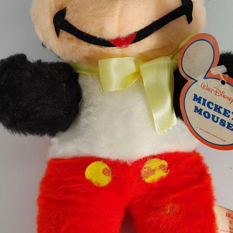 May include: Vintage plush Mickey Mouse toy with a yellow bow tie, red pants with yellow buttons, and a white shirt. The toy has a black face and a white tag that reads "Walt Disney Mickey Mouse" with a copyright symbol and "Walt Disney Productions".
