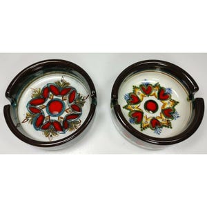 May include: Two ceramic ashtrays with a brown rim and a white center. One ashtray has a red, yellow, and green floral design with a red center. The other ashtray has a red, yellow, and blue heart design with a red center.