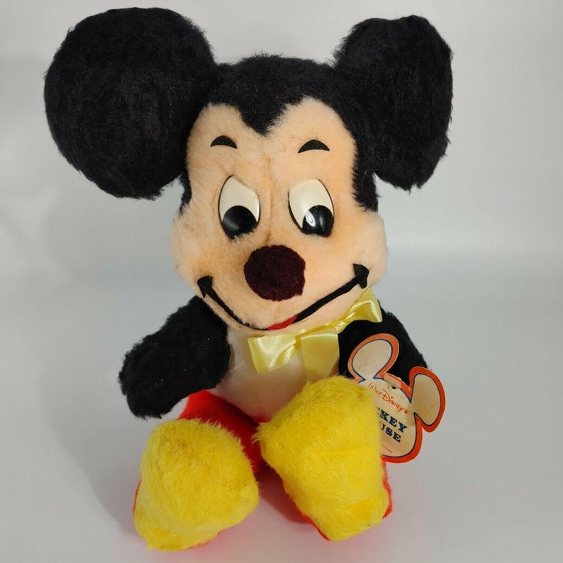 May include: Vintage plush Mickey Mouse toy with black fur, yellow shoes, and a yellow bow tie. The toy is sitting and has a tag that reads "Walt Disney's Mickey Mouse".