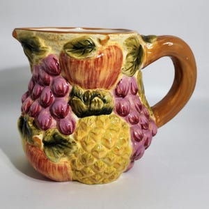 May include: A ceramic pitcher featuring a three-dimensional embossed design of assorted fruits including a yellow pineapple, red apples, and clusters of purple grapes with green leaves. The handle is a solid brown colour with a glossy finish.
