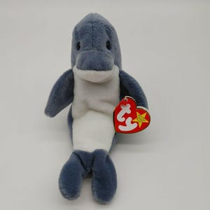 Puede incluir: Un juguete de peluche de delfín azul y blanco Ty Beanie Babies con una etiqueta roja en forma de corazón que dice "Ty Beanie Babies" y tiene una estrella.