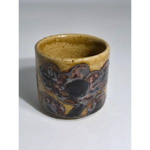 May include: A cylindrical ceramic cup featuring a speckled tan glaze on the interior and rim. The exterior displays a dark, abstract floral pattern in shades of deep brown, black, and muted grey, set against a textured, earthy tan background.