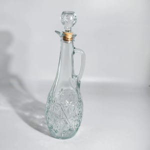 May include: A clear glass cruet bottle featuring a textured diamond and star pattern on the bulbous base. The bottle has a slender neck, a curved side handle, and a removable glass stopper with a cork seal inserted into the top opening.