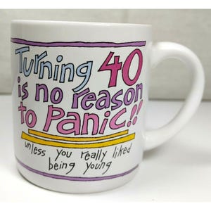 May include: A white ceramic mug featuring the text "Turning 40 is no reason to Panic!! unless you really liked being young" in blue, pink, and black lettering. The design includes two horizontal yellow lines and purple border lines.