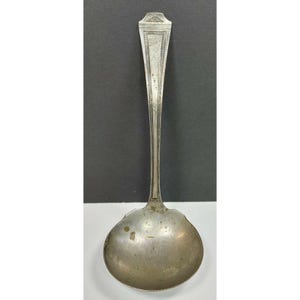 May include: A silver-toned metal ladle featuring a long, slender handle with a decorative etched border pattern. The bowl of the ladle is round and shows signs of age with visible surface patina and minor discoloration throughout the metal finish.
