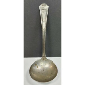 May include: A silver-toned metal soup ladle featuring a long, slender handle with an engraved border pattern and a fan-shaped detail at the top. The bowl of the ladle is round and shows signs of surface wear, including small dark spots and patina.