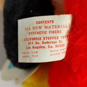 May include: A white tag with red text attached to a red stuffed toy. The tag reads "CONTENTS ALL NEW MATERIAL SYNTHETIC FIBERS CALIFORNIA STUFFED TOYS 611 So. Anderson St. Los Angeles, Ca. 90023 PENN. 173 MASS. T111 OHIO PAI MAINE 56".
