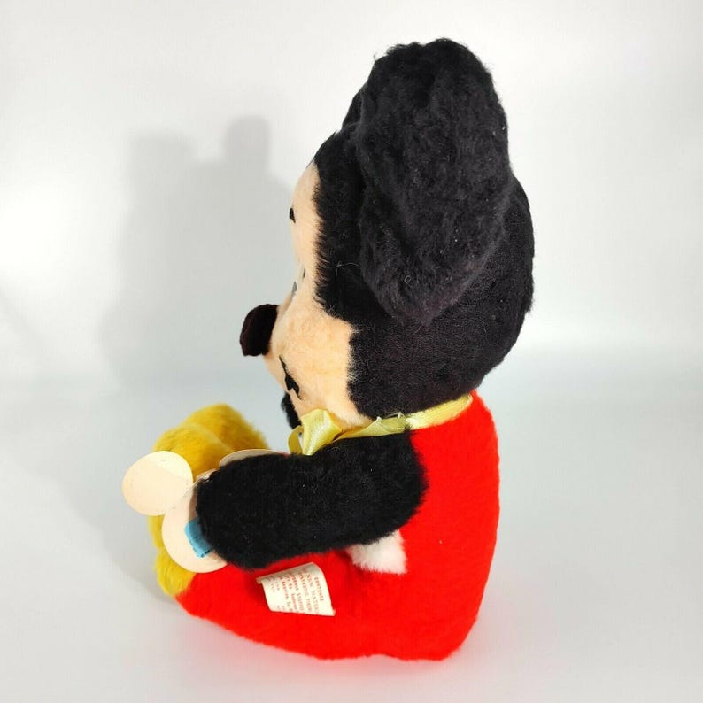 May include: A vintage plush Mickey Mouse toy wearing red shorts and a yellow bow tie. The toy is sitting with its legs crossed and holding a yellow and blue rattle.
