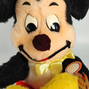 May include: Vintage plush Mickey Mouse toy with black ears, a yellow bow tie, and a red mouth. The toy has a white face with black eyes and a brown nose. The toy is wearing a black and yellow outfit. The toy has a "Disney's" tag attached to it.