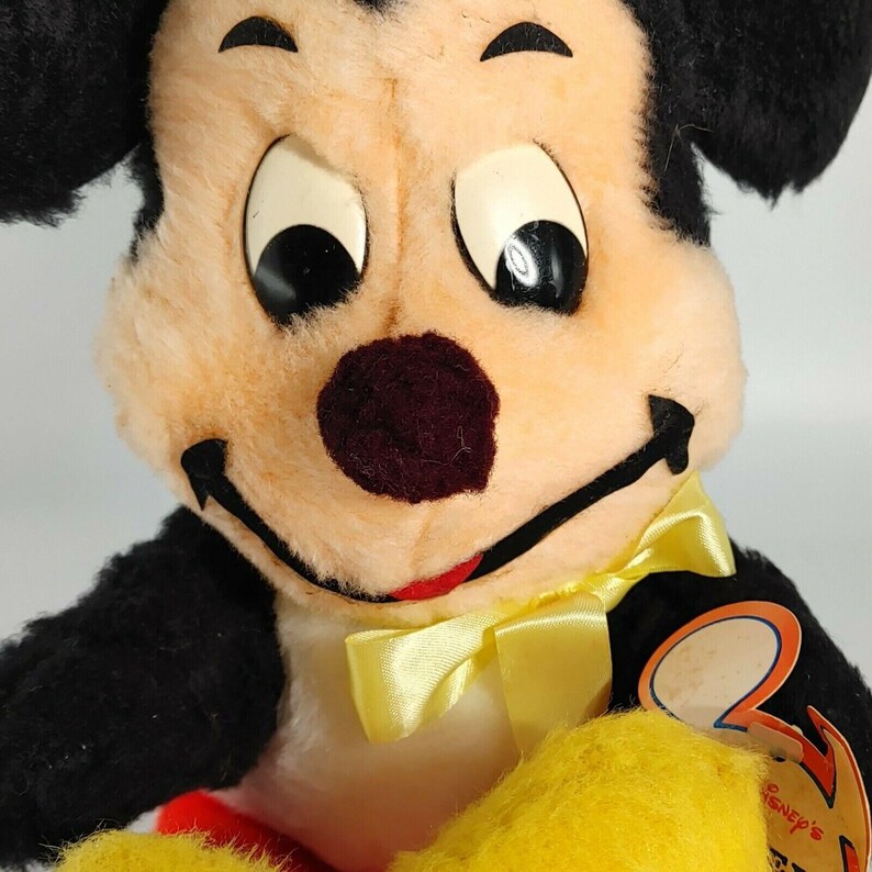 May include: Vintage plush Mickey Mouse toy with black ears, a yellow bow tie, and a red mouth. The toy has a white face with black eyes and a brown nose. The toy is wearing a black and yellow outfit. The toy has a "Disney's" tag attached to it.