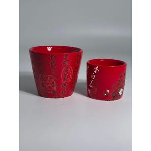 May include: Two red ceramic pots with Asian-inspired calligraphy and floral designs. The larger pot has extensive text, while the smaller one features a floral branch and text. Both pots have a glossy finish.