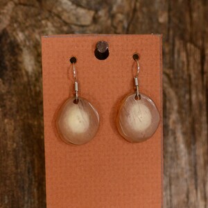 May include: A pair of earrings with a light brown oval-shaped pendant on each earring. The pendants have a translucent, almost milky, appearance. The earrings are displayed on a brown card.