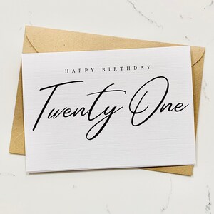 Könnte beinhalten: Eine weiße Karte mit dem Text "Happy Birthday Twenty One" in schwarzer Schrift. Die Karte liegt auf einem braunen Umschlag.
