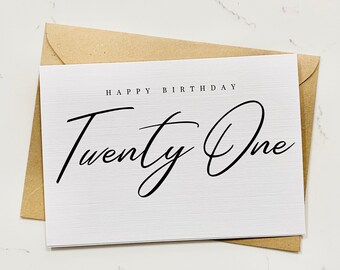 May include: A white card with the text "Happy Birthday Twenty One" in black script font. The card is sitting on a brown envelope.