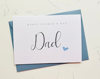 May include: A white card with the text "Happy Father's Day Dad" in black script. A small blue heart is to the right of the word "Dad". The card is on top of a blue envelope.