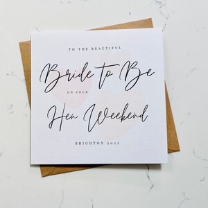K&ouml;nnte beinhalten: Eine wei&szlig;e Karte mit dem Text "To the beautiful Bride to Be on your Hen Weekend Brighton 2022" in schwarzer Schrift. Die Karte hat einen rosa-wei&szlig;en Aquarellhintergrund.