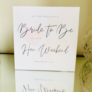 K&ouml;nnte beinhalten: Eine wei&szlig;e Karte mit schwarzer Kalligraphie, auf der "To the beautiful Bride to Be on your Hen Weekend Brighton 2022" steht. Die Karte hat einen rosa Aquarellhintergrund.