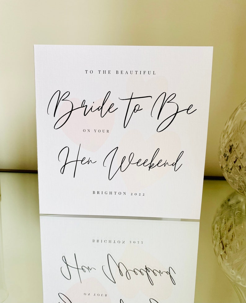 K&ouml;nnte beinhalten: Eine wei&szlig;e Karte mit schwarzer Kalligraphie, auf der "To the beautiful Bride to Be on your Hen Weekend Brighton 2022" steht. Die Karte hat einen rosa Aquarellhintergrund.