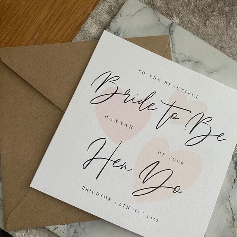 K&ouml;nnte beinhalten: Eine wei&szlig;e Karte mit rosa Aquarellherzen und schwarzer Kalligraphie, auf der steht "To the beautiful Bride to Be Hannah On Your Hen Do Brighton - 6th May 2022".