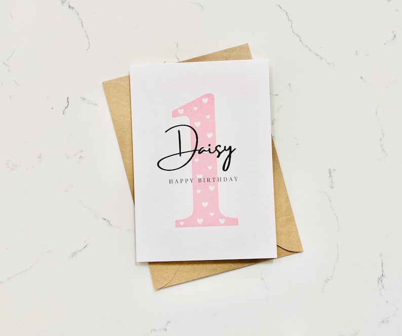 Op de afbeelding: Een roze en witte verjaardagskaart met het nummer &eacute;&eacute;n en de naam Daisy. De kaart zegt "Happy Birthday".
