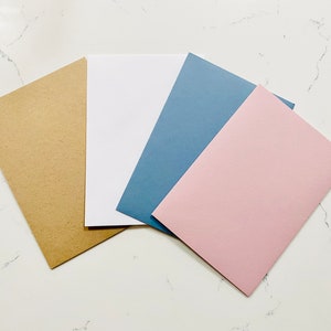 Op de afbeelding: Vier blanco kaarten in verschillende kleuren: bruin, wit, blauw en roze. De kaarten zijn in een waaier op een wit marmeren oppervlak gerangschikt.