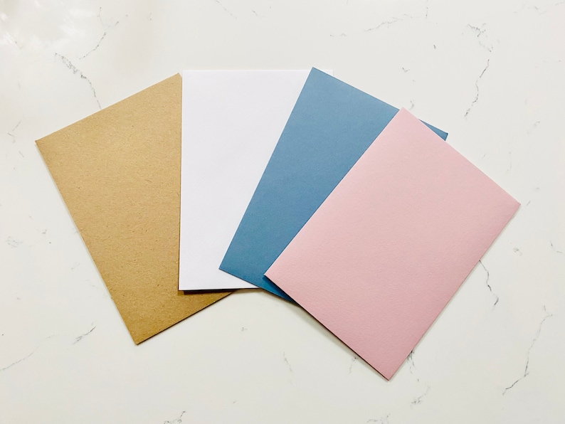 Op de afbeelding: Vier blanco kaarten in verschillende kleuren: bruin, wit, blauw en roze. De kaarten zijn in een waaier op een wit marmeren oppervlak gerangschikt.