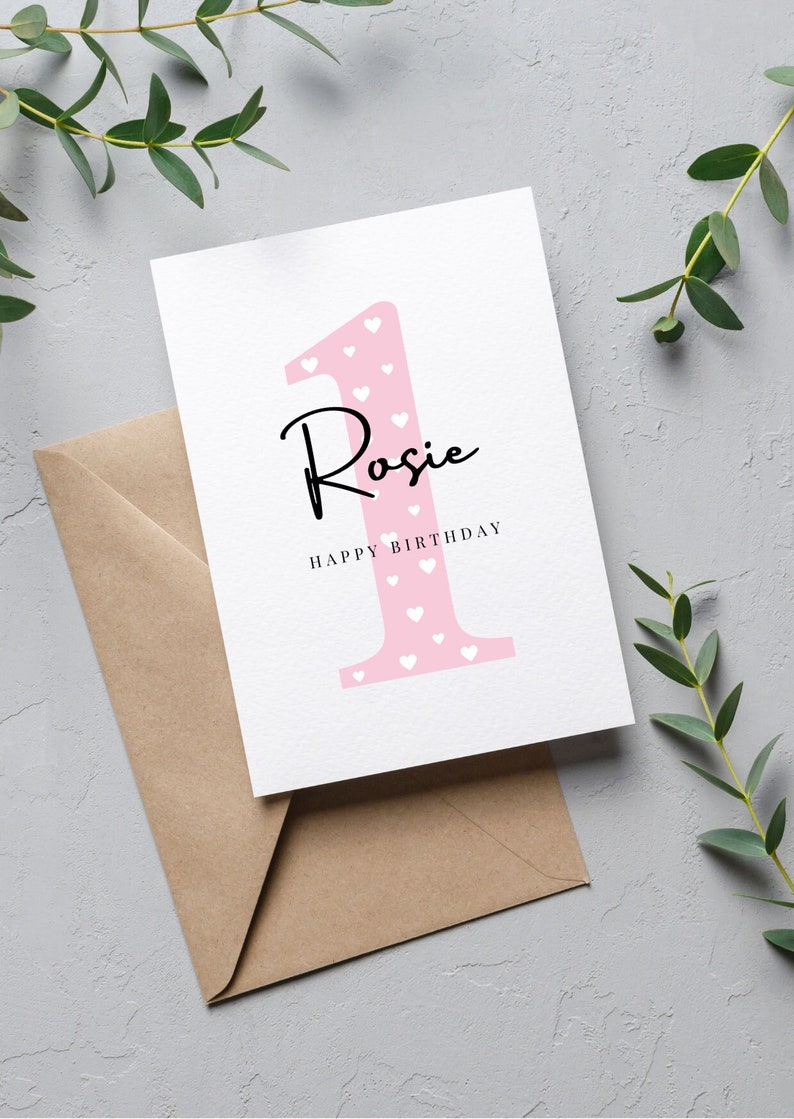 Op de afbeelding: Een roze kaart met een wit nummer &eacute;&eacute;n met witte hartjes erop. De naam "Rosie" is in zwarte cursieve letters onder het nummer &eacute;&eacute;n geschreven. De tekst "Happy Birthday" is in zwart onder de naam geschreven.