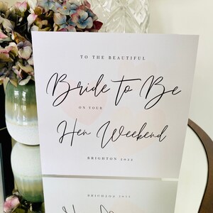 K&ouml;nnte beinhalten: Eine wei&szlig;e Karte mit rosa Aquarell-Akzenten und schwarzem Kalligraphie-Text. Der Text lautet "To the beautiful Bride to Be on your Hen Weekend Brighton 2022".