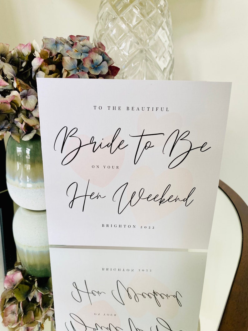 K&ouml;nnte beinhalten: Eine wei&szlig;e Karte mit rosa Aquarell-Akzenten und schwarzem Kalligraphie-Text. Der Text lautet "To the beautiful Bride to Be on your Hen Weekend Brighton 2022".