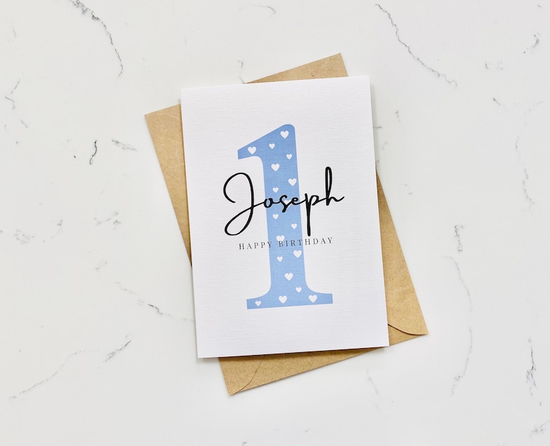 Op de afbeelding: Een witte kaart met een blauw nummer &eacute;&eacute;n met witte hartjes erop. De naam Joseph is in zwarte cursieve letters boven het nummer geschreven. De tekst "Happy Birthday" is in zwart onder het nummer geschreven.