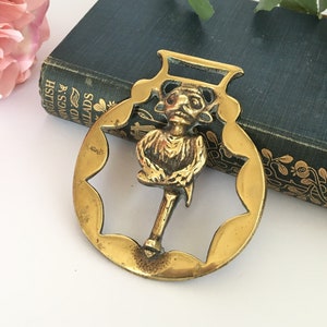 May include: A brass horse brass with a detailed design of a whimsical figure holding a staff. The brass is a warm gold color and has a starburst shape.