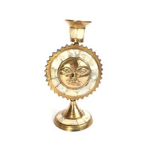 Könnte beinhalten: Ein goldener Kerzenhalter mit einem Sonnen-Gesichtsdesign. Das Sonnen-Gesicht ist aus Perlmutt gefertigt und von einem goldenen Rahmen umgeben. Der Kerzenhalter hat eine runde Basis und einen hohen, schlanken Stiel.