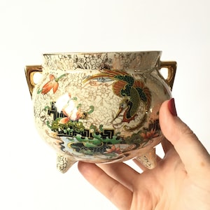 Puede incluir: Una maceta de cerámica blanca con asas doradas y tres patas. La maceta está decorada con un diseño floral y de pájaros en tonos verdes, rojos y dorados.