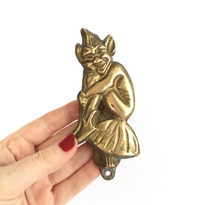 May include: A gold-toned brass door knocker in the shape of a mischievous-looking goblin or imp. The goblin is sitting on a rock or a mushroom with its legs crossed and its hands clasped in front of it.