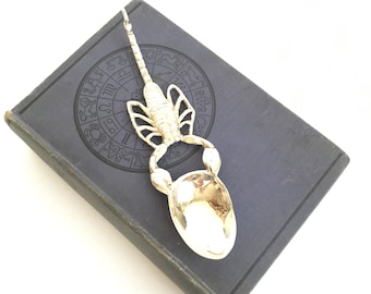 May include: Silver spoon shaped like a scorpion resting on a book with a zodiac wheel design.