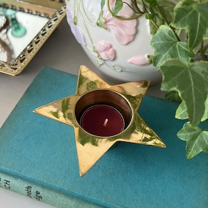 May include: A polished gold-tone metal star-shaped tea light candle holder with a circular center opening containing a small, round, deep red wax candle. The star has five pointed tips and a smooth, reflective surface finish.
