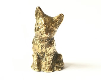 May include: A small, gold-colored metal figurine of a sitting cat. The cat has a detailed, textured surface and is looking down.