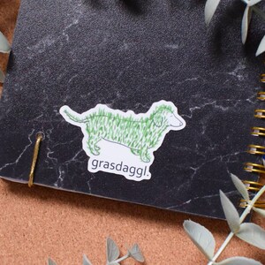 May include: A green dachshund sticker with the text "grassdaggl." on a black marble notebook.