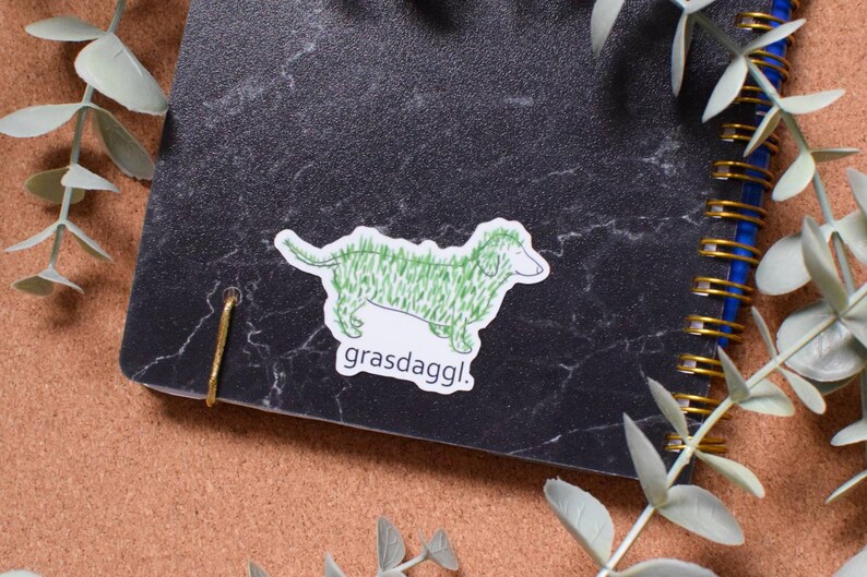 May include: A green dachshund sticker with the text "grassdaggl." on a black marble notebook.