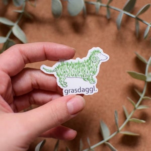 May include: A white sticker with a green line drawing of a dachshund covered in grass. The text "grassdaggl." is below the dog.